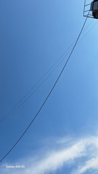 The image depicts a clear blue sky with a few wispy white clouds on the right side. A thin crescent moon is visible high in the sky. Three dark power lines stretch diagonally across the frame from the upper left to the lower right. The top left corner shows a small section of a structure with a metal frame and what appears to be a light-colored roof. The bottom right corner has white text that reads "Galaxy A54 5G". The scene suggests it is daytime, likely late afternoon or early evening given the presence of the moon and the bright sky. The overall mood is serene and vast, emphasizing the expanse of the sky. The location context is Sevan, Armenia.