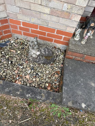 The image depicts a ground-level scene in Minsterley, United Kingdom. The location features a combination of brickwork, concrete slabs, and a gravel-filled area. The brickwork, laid in a running bond pattern, is predominantly red with gray mortar. Adjacent to the brickwork are concrete slabs, varying in texture and color from light gray to darker shades. The gravel area contains small, rounded stones mixed with dried leaves. A weathered stone object sits within the gravel section. Vegetation, including grass and weeds, is present along the edge of a concrete slab. A fragment of a statuette is visible in the upper left corner of the image. No people are visible.