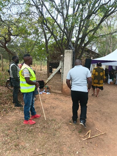 Four people stand outside a gated area. One wears a neon vest and holds a stick, another a mask, and a third a gray shirt. The fourth person, wearing a yellow and black patterned dress, walks toward a tent. A motorcycle is parked nearby. The area appears to be rural, with trees and a simple structure visible through the gate.
