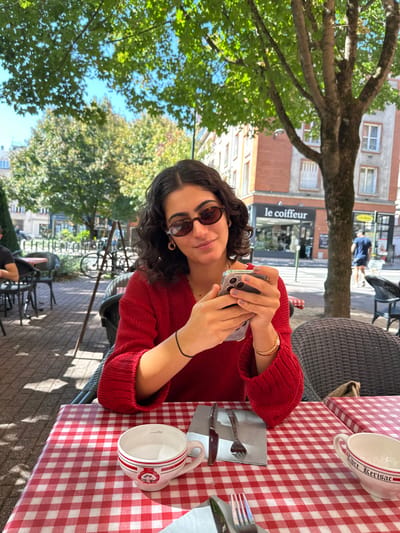 A woman with dark, curly hair, wearing oval-framed sunglasses and a red long-sleeved knitted sweater, is seated at an outdoor cafe table on Rue de la République in Orléans, France. She is holding a smartphone with both hands, positioned over the screen, and looks towards the viewer with a slight smile. Her left wrist has a black elastic band, and her right wrist wears a gold-colored bracelet. Small gold hoop earrings and a thin gold necklace are visible.
The table is covered with a red and white checkered tablecloth. A white ceramic soup bowl with red stripes and a chef insignia, marked with a volume indicator, is on the table to her left. A knife and fork rest on a dark gray napkin beside it. Partially visible to the right is another white ceramic bowl, inscribed "Libre Kerisac."
The outdoor environment features a paved ground with dappled sunlight and shadows from mature green-leafed trees. In the background, a multi-story building displays reddish-brown brickwork combined with lighter-colored facades. The ground floor of this building hosts a business with a black awning reading "le coiffeur" in white text. A person in a blue shirt walks on the sidewalk in the distance. Additional cafe seating, a bicycle rack with a bicycle, and further urban buildings are visible under a clear blue sky.