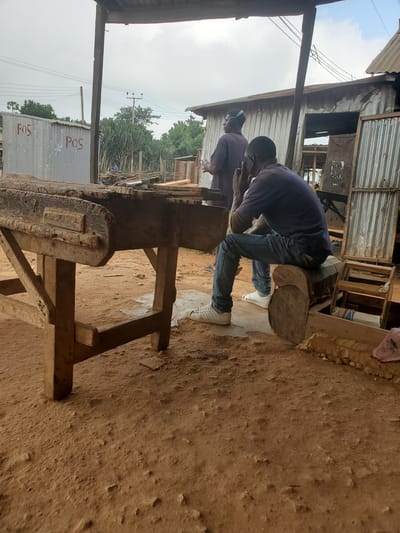 In the city of Barikin Sabon Gidan Kanat, Nigeria, a dusty outdoor workshop setting is depicted. A weathered wooden workbench, supported by sturdy legs, occupies the foreground, its surface bearing marks of past use. Two men are present in the scene. One man, seated on a wooden beam, is facing away from the camera, his right hand near his face as if in thought or on a phone call. He is wearing a dark long-sleeved shirt, blue jeans, and white sneakers. Another man stands behind him, facing left, wearing a dark purple shirt and headphones. To the right of the seated man, a wooden structure with planks and insulation is visible. In the background, corrugated metal structures, some with "POS" written on them, are present along with vegetation and utility poles supporting electrical wires. The ground is covered in dry soil with scattered debris.