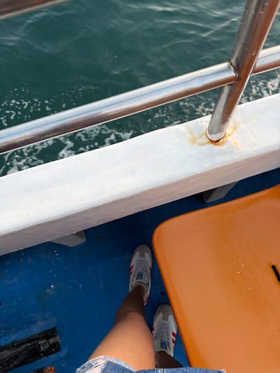 A person's legs, clad in distressed denim shorts and grey and red Adidas sneakers, are visible in the lower half of the frame, standing on a blue deck. To the right, an orange seat or bench occupies a significant portion of the lower right quadrant. The upper half of the image reveals the deep green, rippling water of a body of water, with whitecaps indicating movement. A metal railing runs horizontally across the frame, supported by a vertical post, and a white beam with rust stains runs beneath it. The scene appears to be on a boat, likely in the waters near Pemenang Timur, Indonesia, given the context.