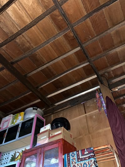 The image depicts an interior view of a rustic structure with a wooden plank ceiling supported by dark wooden beams. The ceiling boards are arranged horizontally and show a natural wood grain. Below the ceiling, various items are stored on shelves and within cabinets.

On the left side, there is a white and pink shelving unit containing boxes, including one with an image of a woman and a blender, and a large silver pot or rice cooker. To the right of this, there is a black helmet and stacked cardboard boxes. One box is labeled "MOREL XARNA".

Further to the right, two colorful umbrellas hang suspended. Next to them, a dark maroon garment, possibly a dress or robe, is draped. Below these items, a wooden cabinet with glass doors reveals stored footwear. In the foreground, a drying rack holds a patterned fabric, possibly a rug or blanket, with maroon, blue, and white designs. The walls appear to be made of unfinished wood panels.

The scene is likely within a residential or storage area, possibly in a rural or less developed setting, given the exposed wooden structure and the collection of household items and storage boxes. The reference to Turumame, Indonesia, suggests this is a specific location within that region. However, there are no explicit visual cues in the image that pinpoint this exact location.