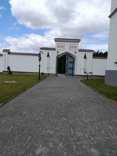 A wide-angle, eye-level shot captures the exterior of the Palace of the Princes of Sapieha in Kosava, Belarus. The foreground is dominated by a grey gravel pathway that curves to the right. To the left of the path is a well-maintained green lawn. The pathway leads towards an open, ornate grey metal gate set within a white archway. The archway is part of a larger white structure with decorative elements. Beyond the gate, a dense forest can be seen. To the left of the pathway and lawn is a white wall with a brown roofline, and a black lamppost stands near the wall. A bench is visible on the grass to the right of the gate. The sky above is a mix of white clouds and blue.