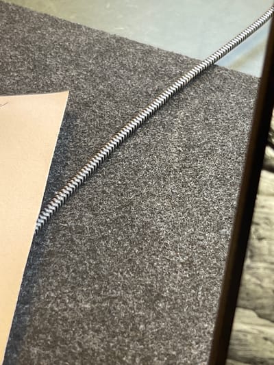 A close-up shot features a grey textured surface, possibly felt or a similar material, partially obscured by a tan-colored rectangular object in the lower portion of the frame. A cord with a black-and-white pattern runs diagonally across the surface. The scene is framed by a dark border at the top. The image provides no information about the location being Kafr Yasif, Israel, or about any people, actions, or interactions.