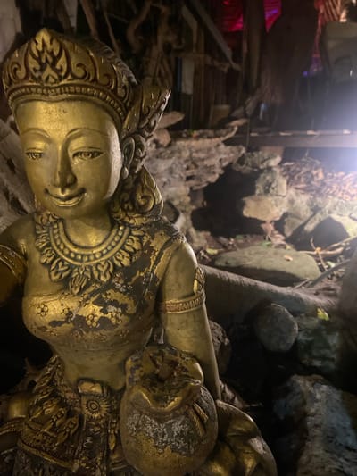 A gilded statue of a reclining Buddha is situated within a cave or temple environment in Baan Don Sai, Thailand. The statue shows signs of age and wear, with areas of corrosion and loss of gilding. The surrounding environment is rocky and includes what appears to be tree roots and cave formations. The area is dimly lit, with some lighting coming from above the scene.