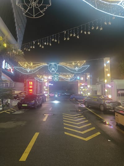 The image depicts a street scene at night in Nanhai District, China, illuminated by decorative lights. Several cars are parked or driving on the wet asphalt road, which is marked with yellow lines and chevrons. A large archway adorned with white string lights and star-shaped ornaments spans the street. On the left, a building facade with illuminated signage is visible, and to the right, a structure with "VE VISION" branding is also lit up. The overall atmosphere suggests a festive or commercial area at night.