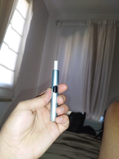 A person's hand is holding a light blue, cylindrical electronic smoking device. The device has a black circular button on its side and a white filter protruding from the top. The background appears to be a bedroom with a large window covered by sheer white curtains on the left and heavier, draped curtains on the right. A dark object, possibly a bag or backpack, is visible on a bed covered with a brown blanket in the background. The lighting suggests it might be daytime, possibly late afternoon, given the soft light filtering through the window. The scene appears to be indoors, likely in a residential setting in Germasogeia Municipality, Cyprus. There is no discernible text in the image. The focus is on the hand and the electronic smoking device.