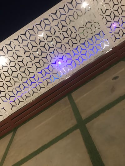 A diagonally-angled view showcases a patterned white panel with geometric, three-dimensional cube-like designs. Behind it, an array of blue lights create a luminous effect. Adjacent to the panel is a brown frame, with a muted beige surface featuring dark green linear patterns in the upper-right corner. The lower-left portion of the image depicts darkness.