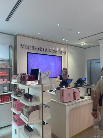 In the image, a Victoria's Secret store in Bergamo, Italy, is depicted, focusing on the checkout area. A female cashier, wearing a black top, is operating a touchscreen point-of-sale system. In front of her are several pink and white striped Victoria's Secret gift boxes and other merchandise. To the right, a person is shown from the chest down, holding a plastic water bottle. Behind the cashier, a large screen displays the Victoria's Secret logo against a purple, sparkly background. The store interior features a clean, modern aesthetic with white and light pink accents, as well as prominent lighting and air conditioning vents on the ceiling. Shelves are visible on the left side, stocked with various beauty products.