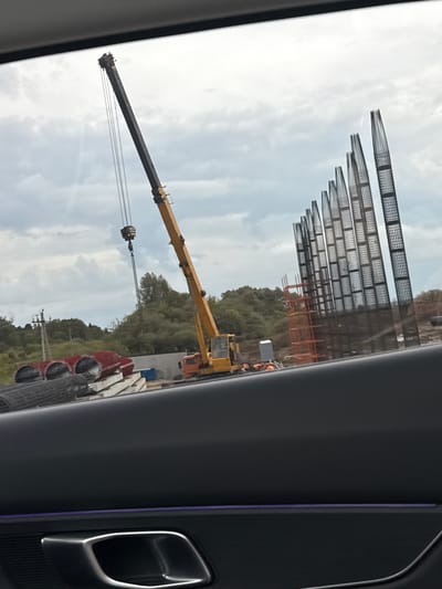 The image captures a construction or industrial site in Kabanikha, Russia, viewed through the window of a vehicle, indicated by the car's dark interior door panel, chrome handle, and a purple ambient light strip visible on the right.

In the background, under a partly cloudy sky, are several large, skeletal industrial structures. These appear to be frameworks, possibly for a large building or facility, composed of numerous vertical and horizontal grid-like sections. To the right of these structures, further construction elements, including some scaffolding, are visible.

In the midground, a prominent yellow and black mobile crane with an extended boom is positioned on the ground, indicating ongoing heavy lifting operations. The crane's hook and cables hang downwards. Scattered around the base of the crane and extending into the foreground are various construction materials, including large red cylindrical objects (possibly pipes or storage containers) and stacks of rectangular, precast concrete or stone blocks, along with rolls of mesh fencing or reinforcement.

Lush green trees and foliage are visible behind the crane and some of the construction materials, suggesting the site is on the edge of a natural area. The time of day appears to be daytime, with soft, diffused light indicating an overcast or partly cloudy sky. No discernible text is visible anywhere in the image. The overall scene depicts an active industrial development or construction project.