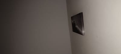A framed picture with an image of a black cat and text is mounted on a light-colored wall. The text on the picture reads: "Nicht alle Engel, manche haben Schnurrhaare." The picture is positioned on the right side of the frame. The background is a plain wall, with a gradient of light and shadow. The location is Winnenden, Germany.