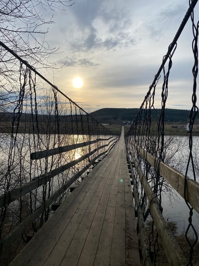 The image depicts a suspension bridge in Pepelyshi, Russia. The bridge is constructed of wooden planks and supported by cables. The environment is rural, with a body of water and forested hills visible in the background. The sun is shining, casting a glow on the scene. There are no people visible in the image. Bare tree branches extend along the sides of the bridge.