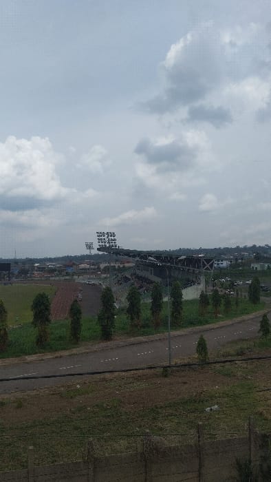 The image shows a view of a football stadium in Buea, Cameroon, with the coordinates 4.163, 9.279. The image was taken from a higher vantage point, looking down at the stadium. The stadium is empty with no people visible, and there are a few trees and bushes growing around it. The sky is overcast with large, puffy clouds. There are a few small buildings in the background, and a road in the foreground with white dashed lines. There is a metal fence in the very foreground. The scene is quiet and peaceful.