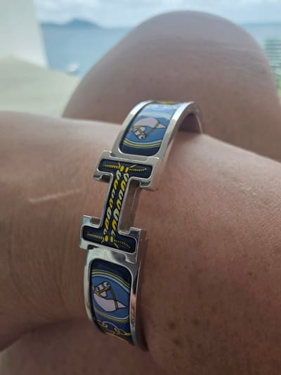 A close-up, eye-level shot shows a person's left arm with a decorative bracelet. The bracelet features a silver "H" logo with intricate yellow and black designs, and the band is adorned with a repeating pattern of stylized fish and nautical elements on a navy blue background. The skin of the arm shows some texture and a faint scar. In the blurred background, water and landmasses can be seen under a cloudy sky, suggesting an outdoor setting possibly near a body of water. This image is not set in Jiyang District, China.