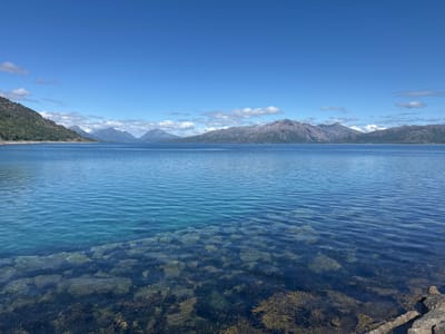 This media file depicts a serene coastal landscape under a clear blue sky with scattered white clouds. The foreground shows the clear, shallow water of a body of water, revealing submerged rocks and seaweed on the seabed. The water exhibits a beautiful gradient of blues and greens, becoming deeper and more opaque further out. In the mid-ground, a vast expanse of calm water stretches towards a distant shoreline. The background is dominated by majestic, rugged mountains, their slopes covered in patches of green vegetation and rocky outcrops. Some of the higher peaks appear to have snow. The overall scene is one of natural tranquility and grandeur. The lighting suggests it is daytime, likely mid-morning or afternoon, given the brightness and the clear visibility of the landscape. The weather appears to be pleasant, with a clear sky and calm water. The location context places this scene in Lødingen, Norway, which is consistent with the mountainous coastal scenery. There is no visible text in the image.