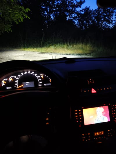The image is taken from inside a car at night, looking forward. The headlights illuminate a dirt or gravel road ahead, bordered by dense vegetation and trees silhouetted against a dark blue sky. The dashboard is visible, with illuminated gauges showing speed (around 50-60 km/h), temperature (23.0°C), and other indicators. The infotainment screen displays a video or image of a person. The location is stated to be Dimitrovche, Bulgaria.