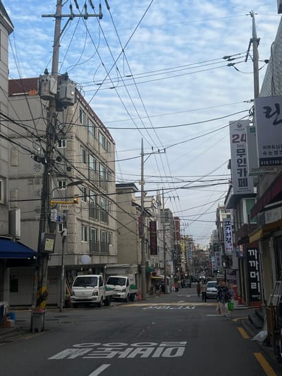 This image depicts an urban street scene in 강서구, South Korea, under a sky with scattered white clouds. The view looks down a street lined by multi-story buildings, characterized by a dense network of overhead utility wires and cables strung between numerous concrete poles.
On the left side of the street, a prominent light-grey building with a stone-textured facade features multiple windows and balconies; Korean text "퐁그렌타운" (Pungnyeon Town) is visible vertically on its side. In front of this building, two white Hyundai Porter II light trucks are parked parallel to the curb. A utility pole on the left is equipped with two transformers and a yellow sign displaying "CCTV" along with Korean text.
The right side of the street is lined with commercial establishments, indicated by numerous vertical and horizontal signs in Korean. Visible signs include "24 hrs 무인카페" (24 hours unmanned cafe) and "고기차림" (Gogi Charim, likely a meat restaurant). The asphalt road surface exhibits white Korean characters painted on it and a yellow dashed line marking the right curb. Several pedestrians are present on the street and sidewalks, with individuals walking in both directions. One pedestrian in a patterned top and light pants walks towards the foreground on the right. Additional vehicles are visible further down the street, which gently slopes uphill into the distance.