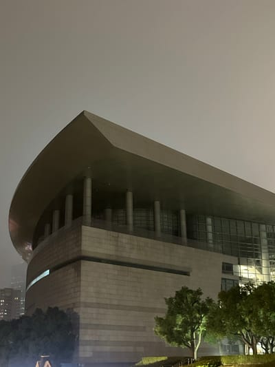 This image depicts a large, modern public building at night, located in the city of Yanqiao, China. The building exhibits a distinctive architectural style, characterized by a prominent, light grey, overhanging roof that curves downwards on the left side, resembling a ship's bow or a bird's wing. This roof is supported by a series of cylindrical columns visible beneath its extensive overhang. The upper facade of the building features large, gridded glass windows, reflecting ambient light from within and around the structure. The lower section of the building is constructed from light-colored, horizontally coursed stone or concrete blocks, with a subtle horizontal recess detail. On the far left, a narrow, horizontal strip of light is visible on a curved segment of the building's lower wall. The sky above is dark and uniformly grey, indicative of night and possibly overcast conditions. In the foreground, several small trees with green foliage are visible, illuminated by external lights, casting a glow on the immediate landscape. No people, specific actions, or interactions are discernible in the scene. Distant, dimly lit buildings are faintly visible on the far left horizon.