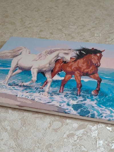 The image shows a painting of two horses running through water at the seashore. One horse is white and the other is brown. The painting is on canvas and appears to be a modern artwork. The background shows a pale blue sky with hints of pink, suggesting a sunset or sunrise. The water is depicted in various shades of blue and white, with splashes and waves. The horses are in motion, with their manes and tails flowing. The scene is dynamic and energetic. The painting is placed on a textured, light-colored surface that appears to be a tablecloth or a similar fabric. The location in Okhtyrka, Ukraine is not depicted in the image.