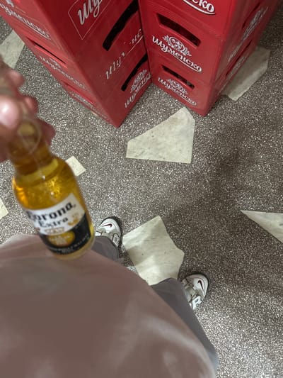 A person is holding a small bottle of Corona Extra beer, with the label clearly visible. The person's hand, arm, and torso are partially in the frame, as are their legs and feet wearing white athletic shoes. The background consists of a tiled floor and stacks of red crates, one of which has "Шуменско" (Shumensko) written on it in white Cyrillic script. The scene appears to be indoors, possibly a storage area or a shop. The location is identified as Gorno Aleksandrovo, Bulgaria.