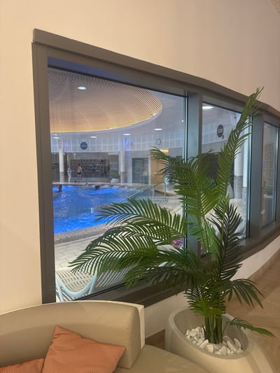 The image shows an interior view through a large, irregularly shaped window. Outside the window is a brightly lit indoor pool area, with people swimming and standing in the water. The pool is blue and is surrounded by architectural elements including a ribbed wall and pillars. Inside, a potted plant with long, green fronds sits to the right of the window, next to a curved sofa with red pillows. The room appears to be well-lit, with the walls a light color. The image location is the city of Marcq-en-Barœul, France.