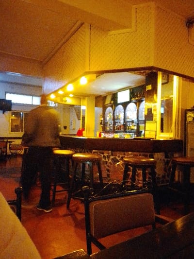 The image depicts the interior of what appears to be a bar or pub, characterized by warm, dim lighting and a cozy atmosphere. The central focus is a well-lit bar counter with a person standing facing it.
The bar itself features a dark, polished wooden countertop. The front of the bar is uniquely decorated with a mosaic or irregular pattern of tiles in earthy tones like brown, white, and black. Several dark wooden bar stools with brown, cushioned seats are lined up along the counter.
Behind the main bar, there's a brightly illuminated back bar area displaying numerous bottles, likely alcoholic beverages, on shelves. This display is framed by what appear to be multiple arched alcoves, adding to the aesthetic. Above the bar counter, a series of bright, warm-toned lights are recessed into an overhang, casting a strong yellow glow on the area.
The overall lighting in the room is warm and yellowish-orange, suggesting an evening setting. The ceiling and upper walls are made of a light brown or beige material with a textured, possibly wicker-like or latticed pattern, featuring architectural angles and slants.
In the foreground and to the left, a person is visible from behind, standing upright with their back to the camera, facing the bar. They are dressed in dark clothing, including what looks like a dark jacket or shirt and dark trousers, and dark shoes with white soles. Their figure is slightly blurred, indicating potential movement or a low-light capture.
To the far left of the image, other tables and chairs can be dimly perceived, hinting at a larger seating area. In the immediate foreground, part of a table and the backrest of a chair with a brown cushion are visible. The floor is a reddish-brown color, possibly tiled or polished.
No discernible text is visible in the image. The overall impression is that of a traditional or classic bar environment designed for relaxation and socializing.