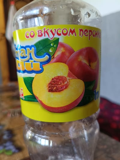 This media file shows a close-up of a clear plastic bottle with a yellow label featuring an image of peaches and text. The label has a bright yellow background. In the foreground, a half of a peach is depicted, showing its vibrant orange flesh and a reddish-brown pit. Behind it, two whole red and yellow peaches are visible, along with green leaves.

The text on the label is in Russian and reads "СО ВКУСОМ персика" which translates to "WITH PEACH FLAVOR". Below this, there is blue and yellow stylized text that appears to be a brand name, possibly "Smart Juice" or similar. On the right side of the label, the number "1л" is visible, indicating a 1-liter volume.

The bottle itself is slightly condensation-covered, suggesting it is a cold beverage. The background is blurred, but hints of a patterned surface (possibly a tablecloth) and dark, indistinct objects are present. The lighting suggests it is daytime. The scene is likely indoors, given the setting cues. No people are visible.
