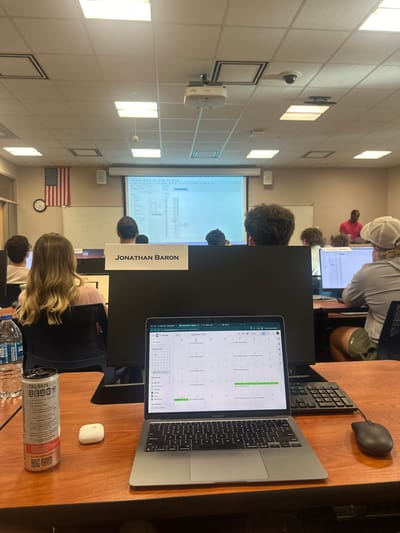 This image depicts a classroom or computer lab located in Fort Myers, United States. The perspective is from a desk in the foreground, looking towards the front of the room.
On the immediate foreground desk, a silver MacBook Air laptop is open, displaying a Google Calendar interface for September 2023. To the right of the laptop, a black computer mouse and a black keyboard are present. To the left of the laptop are a white wireless earbud case, a silver Celsius energy drink can with red accents, and a clear Aquafina water bottle. The desk surface is a polished wood laminate.
Beyond the foreground desk, a large black computer monitor with a white label reading "JONATHAN BARON" is positioned. Several individuals, primarily students, are seated at workstations in rows, facing the front of the room. One student with long, wavy, light-colored hair is seated to the left of the "Jonathan Baron" monitor. Another student wearing a light-colored hat is visible on the far right.
At the front of the room, a large projection screen displays a spreadsheet or data table with columns and rows of information. An Epson projector is mounted on the ceiling above the screen. To the left of the screen, an American flag is mounted on the wall above a circular wall clock. A security camera is affixed to the ceiling in the upper right quadrant of the image. A person in a pink or red shirt is standing on the far right side of the room, partially obscured by students and equipment. The ceiling is a drop ceiling with multiple square fluorescent light fixtures.