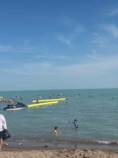 The image depicts a crowded beach scene on a sunny day with a clear blue sky. Numerous people are engaged in various water activities in the turquoise water. In the foreground, a person in a white shirt and dark shorts walks along the sandy shore, carrying a black bag. Two pairs of flip-flops are visible on the sand near the water's edge.
In the water, several individuals are swimming and wading. A black jet ski is positioned to the left, with a person on it. Two long, yellow floating structures are in the water, with people near them. Further out in the water, more people are scattered, some on inflatable devices. Two young boys are in the water, one with a blue and black swimsuit holding a toy water gun, and another boy next to him. The general atmosphere appears to be one of recreational activity and leisure. The location is identified as a beach in the city of Shybarturubek, Kazakhstan.