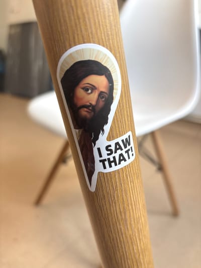 A sticker of Jesus is affixed to a wooden object. The sticker shows a haloed head with dark hair and a beard, and the text "I SAW THAT!". The wooden object is cylindrical and has a light brown wood grain texture. In the background, a white chair is visible with its legs on a light brown floor. The overall scene appears to be indoors.