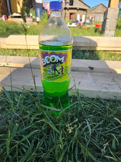 A clear plastic bottle of "BOOM" brand soda, filled with bright green liquid, stands upright on green grass. The bottle has a blue cap and a yellow label featuring cartoon eyes and silhouettes of children playing soccer. In the background, out of focus, are wooden structures, possibly buildings, and other outdoor elements like toy vehicles and scattered lumber. The scene appears to be outdoors, possibly in a rural or suburban setting, suggesting a casual, everyday environment. The image is taken at ground level, looking up towards the bottle.