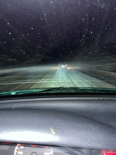 A cracked car windshield with a spiderweb pattern of damage obscuring the view. Headlights of another vehicle are visible through the damaged glass. The interior of the car is visible on the right, including a portion of the dashboard with a speedometer gauge. The windshield's broken edge is adjacent to a textured, dark gray or black interior trim panel. The cracks and debris on the windshield create a distorted and hazardous perspective.