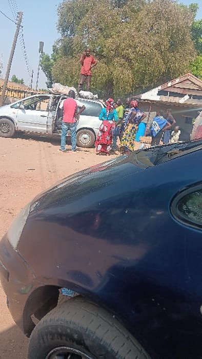 The image depicts an outdoor scene in Langtang, Nigeria, on an unpaved, reddish-brown dirt road under bright daylight. In the midground, a light-colored minivan, likely white or silver, is stationary with its front driver's side door open. Several light-colored, irregularly shaped bundles are secured on its roof rack.
One male individual, wearing a red short-sleeved shirt and dark trousers, stands atop the minivan's roof, positioned among the bundles. Another male, wearing a red t-shirt and blue jeans, stands facing the open driver's side door of the minivan. To the right of the minivan, a group of approximately 5-7 individuals, predominantly women dressed in colorful patterned attire, are gathered. Further in the background, additional individuals are partially visible near a building with a reddish corrugated metal roof. One background figure is identifiable by a blue garment with white text.
The foreground is occupied by the partial view of a dark blue vehicle's front left fender and hood. In the background, a large tree with green-brown foliage dominates the upper right, while utility poles and wires are visible on the upper left.