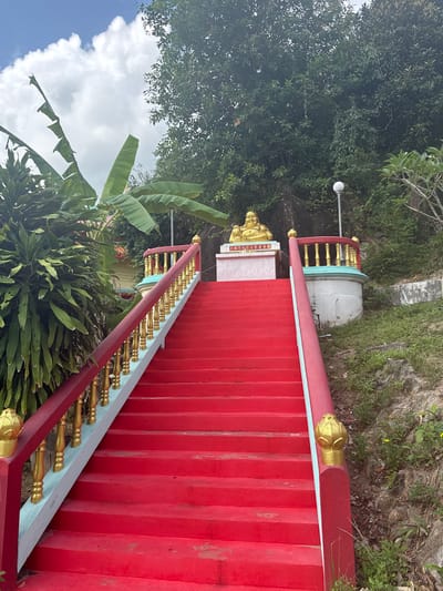 A set of bright red stairs with gilded gold accents leads uphill at a slight angle. The stairs have a red border with gold accents and a railing of red and gold. The steps themselves are painted a consistent, solid red. At the top of the stairs, a gold Buddha statue can be seen. Lush, green vegetation surrounds the stairs on both sides, including banana trees. The sky with scattered clouds is visible through the foliage. The location is Moo 7, Thailand.
