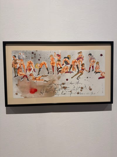 This is a framed artwork hanging on a textured white wall, likely in a gallery or home setting in La Cuesta, Spain. The artwork is a vertical, mixed-media piece featuring a series of illustrated female figures in various contorted and suggestive poses. The figures appear to be rendered in a sketchy, watercolor style, with accents of red, orange, and brown, possibly representing ink or diluted paint. Some of the figures are depicted wearing stockings and other suggestive attire.
Scattered across the background are dark splatters and smudges, along with larger washes of reddish-brown watercolor, giving the impression of stains or abstract elements. To the right of the figures, there is handwritten text in dark ink. The script is cursive and somewhat difficult to decipher, but appears to be French or possibly Spanish. The overall impression of the artwork is one of artistic expression, possibly exploring themes of sensuality or the human form. The lighting in the image suggests it was taken indoors during the daytime, with artificial light casting a shadow to the left of the frame.