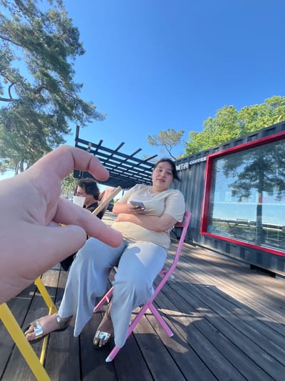 In Ochamchire, Abkhazia, a person's hand is held up in the foreground, forming a heart shape with their fingers. Behind the hand, a woman sits in a pink chair, holding a smartphone. She is wearing a light-colored t-shirt and light-colored pants. To her left, another person sits in a chair, facing away from the camera, holding a white cup. The scene takes place on a wooden deck next to a red-trimmed shipping container with the text "MTA COFFEE" visible. Trees and a clear blue sky are in the background.