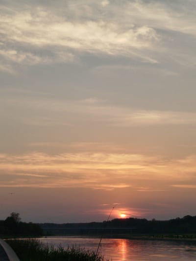This is a photograph of a sunset over a body of water, likely a river, at dusk. The sky is filled with soft, muted colors of orange, pink, and grey, with wispy clouds stretching across. In the foreground, tall grasses and reeds line the bank of the water. A fishing rod is propped up on the bank, extending into the frame. The water reflects the warm hues of the sunset, creating a shimmering effect. Across the water, a silhouetted treeline marks the opposite shore. The overall atmosphere is serene and peaceful, characteristic of a quiet evening in a natural setting. This scene is located in or near Topolina, Poland.