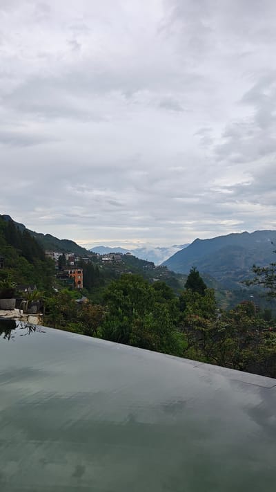 A medium-shot captures an expansive, cloud-laden sky, with the foreground dominated by what appears to be an infinity pool reflecting the sky. The landscape is mountainous with lush green vegetation and scattered buildings, suggesting a populated but natural environment. The location is Cat Cat Village, Vietnam. The horizon line is somewhat obscured by haze or low-lying clouds, adding depth to the scene. A few buildings are visible on the slopes, indicating human presence in the landscape.