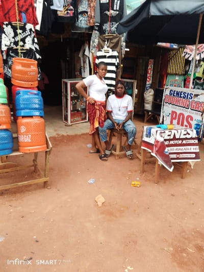 Two people stand outside a small shop with a red dirt ground. One individual sits on a stool, wearing jeans, sandals, and a white t-shirt. The other stands with a red-patterned wrap skirt and a white top. The shop sells clothing and cooking gas, as indicated by the hanging garments and a sign that says "Genesis Cooking Gas". There are also stacked gas cylinders to the left of the pair. The scene appears to be taken during the day.