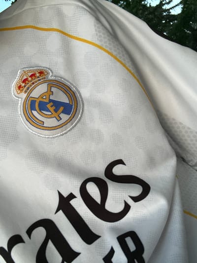 The image shows a close-up of a white Real Madrid jersey with a yellow stripe across the chest and the team's crest. The jersey has a textured pattern of small hexagonal shapes. A portion of the word "Emirates" is visible in large, black, serif font across the lower part of the jersey, indicating it's a sponsor's logo. The background is blurred and appears to be outdoor foliage, suggesting the photo was taken outside. There is no information in the image to suggest a location in Ognianovo, Bulgaria.