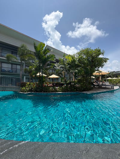 A resort pool area in Bang Muang, Thailand, is captured in this vertical, eye-level shot. The foreground is dominated by a vibrant turquoise swimming pool with rippling water, its edge defined by dark grey tiling. Beyond the pool, a paved area features lounge chairs and umbrellas, some occupied by a few individuals. Lush green foliage and palm trees are interspersed throughout the scene, leading to a multi-story building with balconies. The sky above is a clear blue with scattered white clouds.