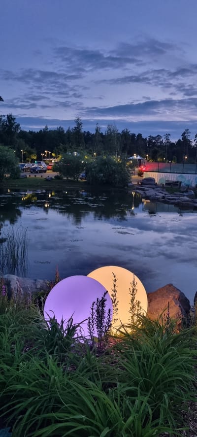 The image depicts an outdoor scene at dusk, likely in Shulgino, Russia. In the foreground, two large, illuminated spherical lamps are situated amongst lush green foliage and rocks. One lamp emits a soft purple glow, while the other shines with a warm yellow light. The lamps are positioned near the edge of a calm body of water, which reflects the dim twilight sky and the surrounding trees.

In the background, across the water, is a well-lit parking lot with several cars. Beyond the parking lot, a dense line of trees forms a dark silhouette against the fading light. Streetlights are visible, illuminating the parking area and the pathway leading into the wooded area. Further in the distance, what appears to be a tennis court or sports facility is illuminated, with a fence enclosing it. The sky above is cloudy, with shades of blue and grey indicating the transition from day to night. The overall impression is of a tranquil, modern park or recreational area transitioning into evening.