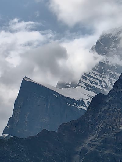 The image shows a dramatic, cloudy sky over a jagged mountain range. The mountains are dark and appear to be made of rock, with patches of snow on their upper slopes and peaks. In the foreground, a sharp, dark mountain face juts upwards, its summit covered in white snow. Behind it, another, more rugged mountain peak is partially obscured by clouds, revealing snow-covered slopes streaked with dark rock. The sky above is filled with a mix of light and dark grey clouds, suggesting an overcast or stormy day. The scene captures the imposing and wild nature of a high-altitude environment. This landscape is consistent with the mountainous region surrounding Gondhla, India, known for its towering peaks and dramatic weather patterns.