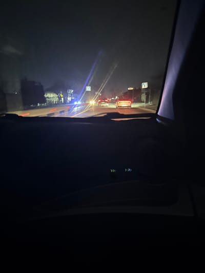 The image shows a dark interior view from a vehicle at night. Outside the window, there's a street with traffic, street lights, and buildings. The reflections and light streaks suggest a wet or foggy atmosphere. Inside the vehicle, the interior is mostly dark, obscuring details, but a headrest is partially visible.