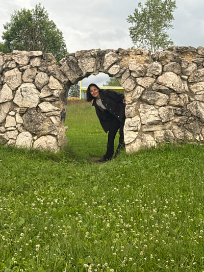 A person is peeking through a stone archway in Chusovoy, Russia. The archway is constructed of rough, irregular stones and appears to be part of a larger structure or ruin. The person, a woman with dark hair, is wearing a dark jacket over a light-colored shirt and dark pants. She is smiling and looking towards the camera. The area in front of the archway is covered in lush green grass with small white flowers. In the background, beyond the archway, is more green grass and trees under a cloudy sky. There is a hint of a yellow structure in the far background.