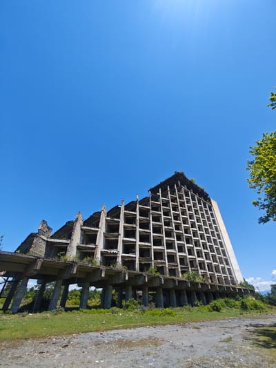 A low-angle, wide-shot photograph captures a large, dilapidated concrete building under a bright, clear blue sky. The multi-story structure, possibly a former hotel or resort, is severely damaged, with much of its exterior facade missing, exposing a grid-like framework of concrete beams and columns. Small plants and overgrown vegetation sprout from the building's ledges and upper sections, indicating prolonged abandonment.

The building stands on a series of substantial concrete pillars, beneath which more wild greenery and bushes grow. The foreground features an unpaved, uneven ground covered with dirt, gravel, and patches of grass. To the right, a portion of a lush green tree frames the scene, contrasting with the stark concrete. The day is sunny, evident from the deep blue sky, bright illumination, and a sun flare visible in the upper-left corner. The overall impression is one of decay, neglect, and nature reclaiming a once-grand structure. No people or visible text are present in the image. This scene is located in Warcha, Abkhazia.