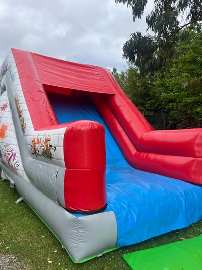 This media file shows a large, inflatable play structure in an outdoor setting. The structure is primarily red and blue, with some grey sections that feature colorful graffiti-style artwork. The artwork includes stylized text and cartoonish characters. The inflatable appears to be situated on a grassy area, with trees and foliage visible in the background. A portion of a green inflatable or barrier is also visible to the right of the main structure. The ground is covered in green grass with some small white flowers. The sky is overcast, suggesting a cloudy or possibly rainy day. The image is taken from a high angle, looking down and across the inflatable. There is no clear indication of people or a specific activity taking place, other than the presence of the play structure itself, which is typically used for recreation. The overall impression is of an outdoor event or setup for children's entertainment. The location context provided is Kilpedder, Ireland, which implies the scene is set in that region.