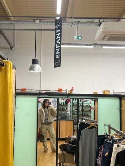 The image captures a female individual taking a mirror selfie inside a retail establishment located in Vendenheim, France. The subject, with dark, medium-length hair and eyeglasses, is wearing a light beige faux fur jacket over a grey top, light khaki cargo pants, and tan platform shoes. She holds a dark brown patterned handbag in her left hand and a smartphone in her right, positioning it for the photograph.

The mirror is set into a wall section painted a pale mint green, bordered by a dark frame. To the immediate left of the mirror, a yellow fabric curtain or partition is visible. Above the mirror, the wall is white, featuring a shelf with small orange labels and several wooden boxes. A red fire extinguisher sign is also affixed to this upper wall section.

The ceiling above is exposed, revealing metal conduit trays, multiple fluorescent strip lights, and a single suspended industrial-style lamp with a metal shade. A black banner hangs from the ceiling, displaying "ESPACE ENFANT" (French for "CHILD SPACE") with a left-pointing arrow. Additionally, a green exit sign with a running man icon and a right-pointing arrow, along with an air conditioning unit, are visible suspended from the ceiling.

The reflection in the mirror shows a portion of the store's interior. Behind the subject, a wooden counter area with multiple shelves displays numerous pairs of shoes. To the right of this counter, an upholstered armchair with dark fabric draped over it is visible. Further to the right in the reflection, clothing racks are present, holding various garments including a grey long-sleeved shirt and denim items.
