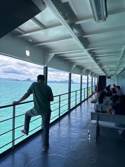 The image depicts the interior of a ferry boat, likely en route to or from Thong Nian, Thailand, given the tropical island scenery visible through the large windows. The ferry's deck is painted a dark color and appears wet in places. A metal railing runs along the windows, separating passengers from the turquoise water outside.

In the foreground, a man stands facing away from the camera, leaning on the railing. He is wearing a green polo shirt, dark pants, and purple sneakers, with his right leg bent and resting on the railing.

Further inside the ferry, several passengers are seated on benches. The benches appear to be made of metal and are arranged in rows. In the middle ground, a person is partially visible seated on a bench, looking towards the camera. A bottle of water rests on the floor near the benches.

The interior of the ferry features a white ceiling with exposed beams and fluorescent lighting fixtures. A staircase is visible in the background, leading to another level of the ferry.

The view outside the windows reveals a scenic panorama of islands with distinctive mountain formations against a cloudy sky. The water is a vibrant turquoise color. The scene suggests a relaxed journey through a picturesque coastal region.